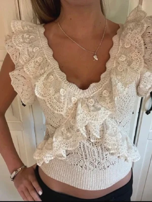Vit spetsblus med volangdetaljer - Superfin vit blus i spets med breda volangdetaljer runt axlar och halsringning. Toppen är ärmlös och har en v-ringad rygg. Perfekt för dig som gillar romantisk och feminin stil. Materialet är stickat och känns luftigt och lätt.