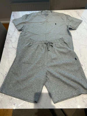 Grå pyjamas-set med shorts - Säljer ett grått pyjamas-set från Polo Ralph Lauren med t-shirt och shorts. Setet är gjort i mjukt bomullsmaterial och har den klassiska Polo-loggan broderad på både tröjan och shortsen. Shortsen har dragsko i midjan och sidofickor. Perfekt för chill dagar hemma.