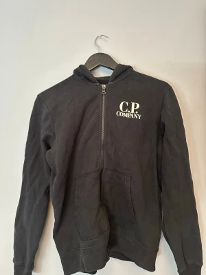 Svart hoodie från C.P. Company - Pris kan diskuteras! Svart hoodie med dragkedja och de klassiska goggles från C.P. Company. Trycket har lossnat lite från bröstet men inget man tänker på vid användning. Storleken är 14Y men sitter som en S/M. 