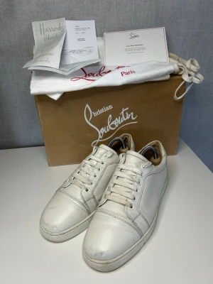Vita sneakers från Christian Louboutin - Stilrena vita sneakers från Christian Louboutin med klassisk låg siluett och vita snören. Skorna har en minimalistisk design med diskreta sömmar och är tillverkade i slätt läder. Perfekta för dig som gillar exklusiva märken och vill ha en clean look. Saknar innersulor så man kan behöva lägga dit egna. Allt og inklusive kvitto, behöver en tvätt! Lägg märke till hål i hälen 