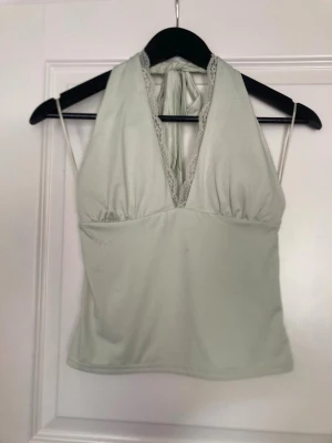 Ljusgrön halterneck linnetopp - Snygg ljusgrön halterneck topp med djup v-ringning och spetsdetaljer längs urringningen. Toppen knyts i nacken och har en ärmlös design som ger en fräsch och trendig look. Perfekt för varma dagar eller sommarens alla tillfällen.