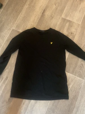 Svart långärmad tröja med gul logga - En stilren svart långärmad tröja med rund hals och en liten gul broderad logga på bröstet. Tröjan har en enkel och clean design som passar till det mesta. Perfekt för dig som gillar minimalistisk stil.