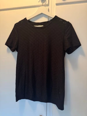 Svart spetsmönstrad t-shirt - Svart t-shirt med korta ärmar och spetsmönster över hela plagget. T-shirten har rund halsringning och är tillverkad i ett mjukt tyg som känns bekvämt mot huden. Perfekt för dig som vill ha en basic look med en twist.