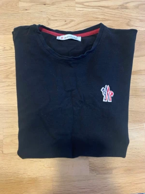 Svart Moncler t-shirt med logga - Snygg mörkblå t-shirt från Moncler med klassisk rund hals och korta ärmar. På bröstet sitter Monclers ikoniska logga i vitt, rött och blått. T-shirten har en enkel och stilren design med diskreta detaljer och är tillverkad i mjuk bomull.