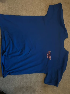 Gallery Dept  t-shirt - Snygg blå t-shirt från Gallery Dept. med orange tryck fram och stort tryck på ryggen där det står 'GALLERY DEPT. HOLLYWOOD, CA.'. Klassisk passform och korta ärmar, perfekt till en avslappnad stil. Kvaliteten är riktigt bra