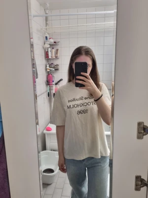 Beige t-shirt Acne Studios Stockholm - Säljer en beige t-shirt från Acne Studios med tryck 'STOCKHOLM 1996' på bröstet. T-shirten har en avslappnad passform, rund hals och korta ärmar. Perfekt till jeans eller shorts för en enkel och clean look.