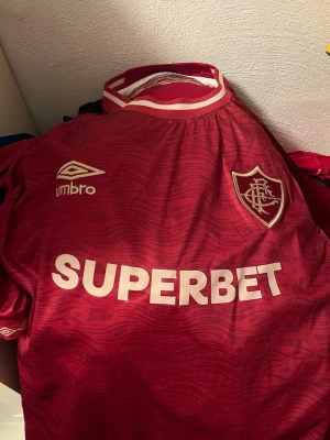 Nudie jeans - Säljer en röd fotbollströja från Umbro med vit logga och klubbmärke på bröstet samt texten SUPERBET tryckt över magen. Tröjan har korta ärmar, rund hals med vit och röd kant och ett subtilt mönster i tyget. Perfekt för match eller träning.