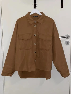 Brun overshirt med fickor - Snygg brun overshirt med klassisk krage och stora bröstfickor med lock. Rak och avslappnad passform, knäpps med knappar framtill. Perfekt lager-på-lager-plagg i mjukt material. Passar till många olika stilar.