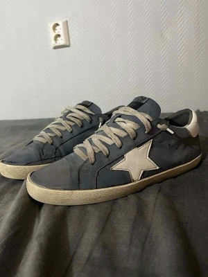 Golden goose  - Snygga svarta sneakers från Golden Goose med ikonisk vit stjärna på sidan och vit häl. Skorna har beige snören och slitna detaljer för en cool, avslappnad look. Tillverkade i Italien och har en klassisk låg profil.