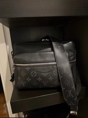 Louis vuttion Svart axelväska med monogram - Snygg svart axelväska från Louis Vuitton med klassiskt monogrammönster i grått. Väskan har justerbar axelrem, silverfärgade dragkedjor och flera fack. Tillverkad i tåligt lädermaterial och har en stilren, boxig form.