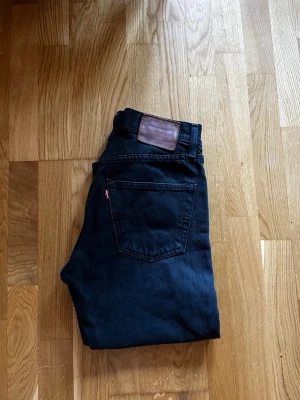 Svarta raka jeans från Levi's - Säljer ett par svarta Levi's jeans med klassisk femficksdesign och rak passform. Jeansen har en brun läderpatch bak i midjan och den ikoniska röda Levi's-taggen på bakfickan och även knappar.  Perfekta till en avslappnad streetstyle-look. Innerbenslängd: 77cm. Midja: 36cm. 