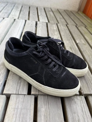 Svarta sneakers från Axel Arigato - Stilrena svarta sneakers från Axel Arigato i mocka med vita sulor och svarta skosnören. Diskret logga på sidan och klassisk låg modell som passar till det mesta. Perfekta för dig som gillar minimalistisk och clean stil.