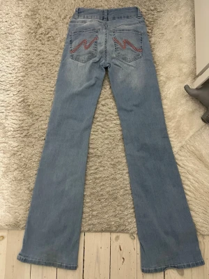 Ljusblå bootcut jeans med brodyr - Säljer ett par ljusblå bootcut jeans med broderade detaljer i rött och vitt på bakfickorna. Jeansen har låg midja, dubbla knappar framtill och klassisk femficksdesign. Materialet är mjukt denim i bomull med lätt stretch för extra komfort.