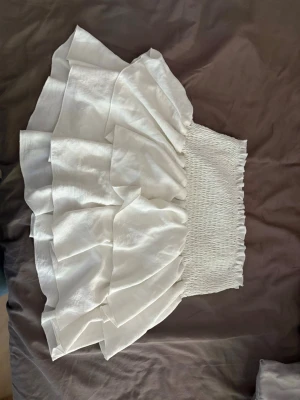 Vit volangkjol med smock - Supersöt vit kjol med bred smock i midjan och flera lager volanger. Kjolen är kort och har en luftig känsla, perfekt för varma dagar. Passar till både sneakers och sandaler för en chill sommarlook.