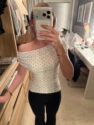 Vit offshoulder topp med prickar - Säljer en vit offshoulder topp med svarta prickar. Toppen är tight och har en snygg, figurnära passform. Materialet känns mjukt och stretchigt, perfekt för dig som gillar en trendig och stilren look.