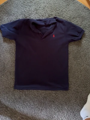 Mörkblå pikétröja från Polo Ralph Lauren - Klassisk mörkblå pikétröja från Polo Ralph Lauren med röd broderad logga på bröstet. Tröjan har korta ärmar, krage och knappar vid halsen. Perfekt till jeans eller shorts för en clean och stilren look.