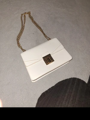 Vit axelväska med guldkedja - Snygg vit axelväska i rektangulär form med guldfärgad kedjerem och elegant guldfärgat spänne framtill. Väskan har stilren design och är tillverkad i slätt material som ger en lyxig känsla. Perfekt för dig som gillar minimalistisk och trendig stil.