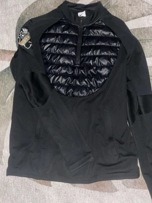 Svart Nike tröja med AIK-märke - Svart långärmad tröja från Nike med AIK-märke på ärmen. Framsidan har ett vadderat parti med quiltad design och halv dragkedja. Tröjan har hög krage och fickor framtill. Perfekt för dig som gillar sportig stil och AIK.