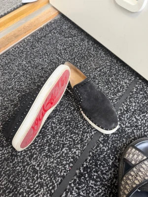 Svarta loafers med nitar och röd sula - Säljer ett par svarta loafers i mocka med vita sulor och coola nitar runt hela skon. Sulan har en röd detalj undertill. Skorna har en stilren slip-on design och passar perfekt för dig som vill sticka ut lite extra.