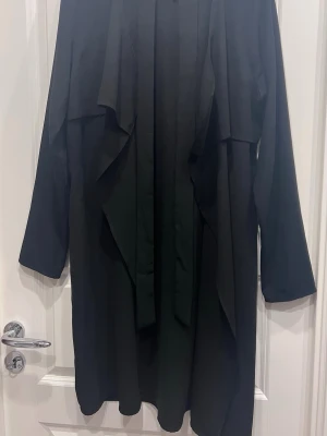 Svart tunn trenchcoat med skärp. - Stilren svart tunn trenchcoat med långa ärmar och löst fall. Jackan har ett matchande skärp i midjan och en öppen front med breda slag. Materialet ser ut att vara tunt och mjukt, perfekt för lager på lager. Enkel och modern design som passar till det mesta.