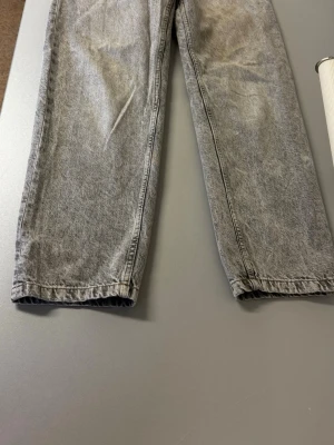 Grå tvättade jeans från Bershka - Säljer ett par grå tvättade jeans från Bershka med raka ben och klassisk femficksmodell. Jeansen har metallknapp, dragkedja och detaljer i kopparfärg. Materialet är denim i bomull. Perfekta för en avslappnad och trendig look. De är välanvända.