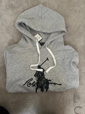 Grå hoodie från Polo Ralph Lauren - Snygg grå hoodie med stor svart Polo Ralph Lauren-logga och polospelare på bröstet. Hoodien har vit dragsko i huvan och en klassisk känguruficka framtill. Tillverkad i mjukt sweatshirtmaterial, perfekt för en chill och avslappnad stil.Priset går att diskutera vid snabb affär, finn de frågor så är de bara o skriv 😊