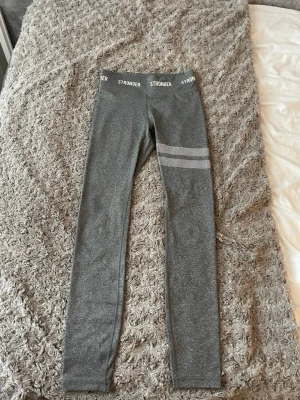 Grå träningstights från Stronger - Snygga grå leggings från Stronger med vita logotyper i midjan och två vita randdetaljer på ena benet. Stretchigt material som sitter tight och bekvämt. Perfekta för träning eller chill dagar.
