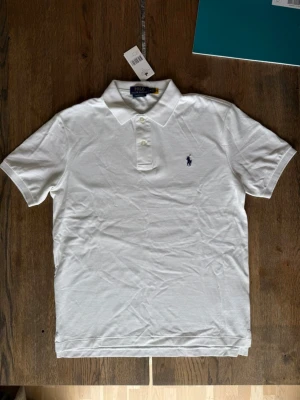 Vit pikétröja från Polo Ralph Lauren - Klassisk vit pikétröja från Polo Ralph Lauren med korta ärmar och krage. Tröjan har två knappar framtill och den ikoniska broderade loggan på bröstet. Perfekt till jeans eller shorts för en clean och stilren look.