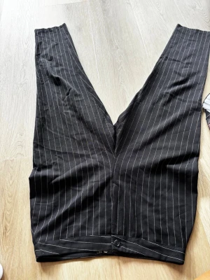 Svarta kostymbyxor med kritstreck - Snygga svarta kostymbyxor med vita kritstrecksränder. Byxorna har rak passform och klassisk midja med knappstängning. Perfekta för en stilren och trendig look. Materialet ser ut att vara en mjuk och lätt tygblandning.