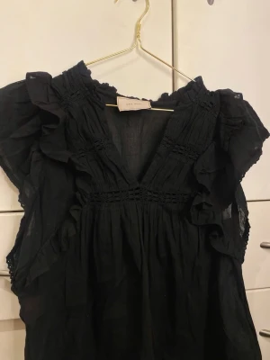 Svart neo noir bluse - Pen svart bluse med rysjedetaljer og V-hals. Blusen har korte ermer og er laget av et lett, luftig materiale som gir en avslappet, men elegant stil. Perfekt å kombinere med jeans eller skjørt for et trendy antrekk.