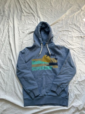 Blå hoodie från Superdry med tryck - Säljer en blå hoodie från Superdry med ett färgglatt tryck av solnedgång, palmer och vågor på bröstet. Hoodien har vit dragsko i huvan, ribbade muddar och en stor magficka. Perfekt för chill dagar och streetwear vibes.