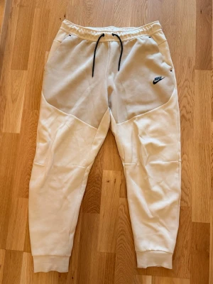 Nike beige mjukisbyxor med snörning - Säljer ett par beige mjukisbyxor från Nike med ljusgrå panel framtill, svarta snören i midjan och ribbade muddar vid benslut. Byxorna har fickor fram och en svart dragkedja bak. Perfekta för chill dagar eller träning.