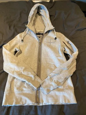 Ljusgrå zip-hoodie från Pelle P - Snygg ljusgrå hoodie från Pelle P med dragkedja framtill, justerbar huva med snören och broderad logga på bröstet. Hoodien har långa ärmar, fickor på sidorna och är tillverkad i mjukt sweatshirtmaterial. Perfekt för en avslappnad och sportig stil.
