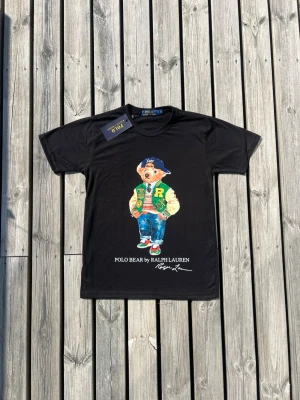 Svart Polo Bear t-shirt från Ralph Lauren - Svart t-shirt med Polo Bear-motiv från Ralph Lauren. Trycket visar en björn i färgglada kläder och texten 'POLO BEAR BY RALPH LAUREN' under. Klassisk passform med rund hals och korta ärmar. Perfekt för dig som gillar streetstyle och ikoniska prints.
