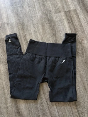 Svarta seamless leggings Gymshark - Snygga svarta seamless leggings från Gymshark med hög midja och diskret mönster längs benen. Stretchigt material som sitter tight och bekvämt. Perfekta för träning eller chill. Gymshark-logga på låret och vristen för en sportig look.