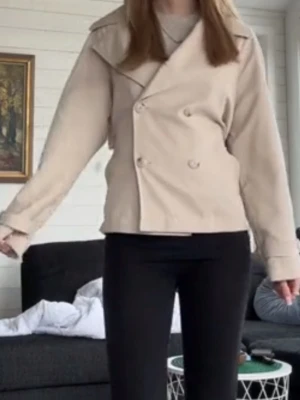 Beige kort trenchcoat med knytband - Stilren beige trenchcoat med dubbelknäppning framtill och brett knytband i midjan bak. Jackan har klassiska axelklaffar och en kortare, boxig passform. Perfekt för dig som vill ha en trendig och enkel look. Materialet ser ut att vara mjukt tyg, troligen bomullsblandning. Bilderna är från min TikTok så om du vill ha nyare så skriv till mig så fixar jag!
