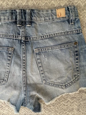 Blå jeansshorts med slitningar - Säljer ett par blå jeansshorts från Ü i storlek 152. Shortsen har slitna detaljer, fransig kant och klassisk femficksdesign. Perfekta för varma dagar och ger en avslappnad look.