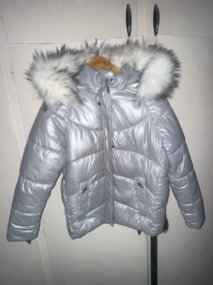 Silver pufferjacka med fuskpäls - Snygg silverfärgad pufferjacka med stor huva och vitgrå fuskpäls. Jackan har dragkedja, tryckknappar och två fickor framtill. Materialet är glansigt och quiltat för extra värme. Perfekt för kalla vinterdagar och ger en cool look.