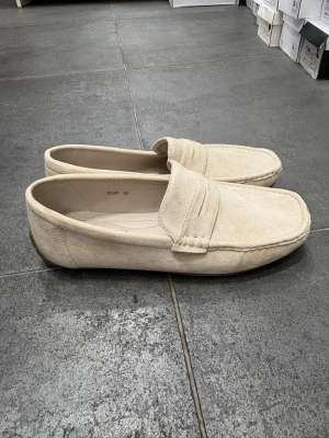 Beige mockaloafers  - Snygga beige loafers  i mjuk mocka. Skorna har klassisk pennyloafers-design med rundad tå och diskret söm längs kanten. Yttersulan är i gummi för extra komfort och grepp. Perfekta för en stilren och avslappnad look.