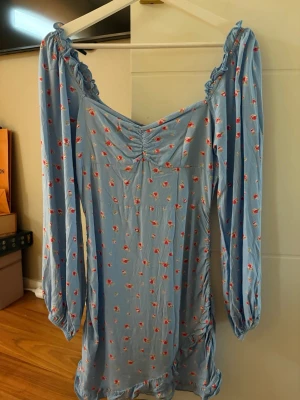 Blommig blå långärmad klänning - Söt ljusblå klänning med små röda blommor, långa puffärmar och rynkade detaljer vid bysten. Klänningen har volangkant nertill och axelband med volang. Perfekt för en somrig look.