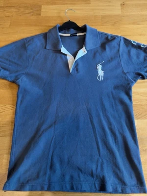 Ralph lauren pike - Klassisk marinblå pikétröja med vit krage och knappar. Tröjan har ett stort vitt broderat polospelar-motiv på bröstet. Perfekt till jeans eller shorts för en avslappnad men snygg look.
