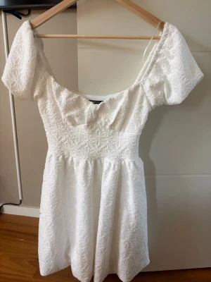 Vit kort klänning med puffärm - Söt vit kort klänning med puffärmar och broderat blommönster. Klänningen har en v-ringad urringning och markerad midja som ger en feminin siluett. Perfekt för sommarens alla tillfällen.