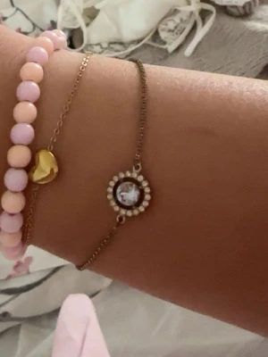 silvrigt  armband med pärldetaljer - Snyggt armband i silvrig  metall med en rund dekoration i mitten. Dekorationen har en stor klar sten i mitten omgiven av små vita pärlor. Tunt kedjeband med karbinlås. Perfekt för dig som gillar stilrena och eleganta smycken.
