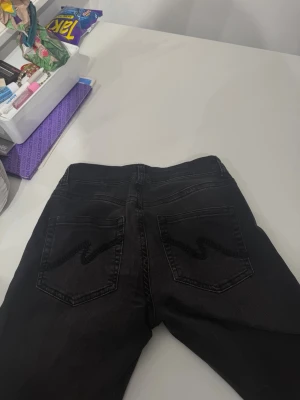 Svarta jeans med slitningar - Säljer ett par svarta jeans med raka ben och slitningar vid knäna. Jeansen har klassiska bakfickor med zigzag-detalj av märket ”Nelly”  och är tillverkade i ett mjukt denimtyg. Perfekta för en avslappnad och trendig look.