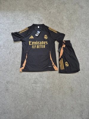 Real Madrid x Adidas - Snyggt svart träningsset från Adidas med Real Madrid-logga och guldiga detaljer. Tröjan har korta ärmar, rund hals och texten 'Emirates Fly Better' på bröstet. Matchande shorts med guldiga ränder och klubbmärke. Perfekt för fotbollsträning eller gymmet.