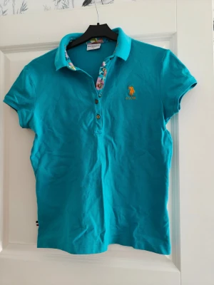 Turkos pikétröja från U.S. Polo Assn. - Snygg turkos pikétröja från U.S. Polo Assn. med korta ärmar och färgglada detaljer på insidan av kragen och knappslån. Tröjan har broderad logga i gult på bröstet och klassisk krage med tre knappar. Perfekt för en fräsch och sportig look.