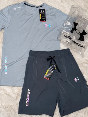 Under Armour träningsset i grått - Snyggt träningsset från Under Armour med en ljusgrå t-shirt och mörkgrå shorts. Båda plaggen har holografiska loggor och textdetaljer. Tillverkade i ett lätt och snabbtorkande material som passar perfekt för träning.