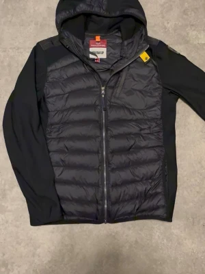 Svart dunjacka med huva från Parajumpers - Snygg svart dunjacka från Parajumpers med huva och dragkedja framtill. Jackan har quiltad front, släta ärmar och axelpartier samt en gul detalj vid kragen. Märkespatch på ärmen och innerfoder med 'WARM UP'-etikett.