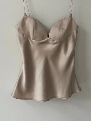 Champagnefärgad satinlinnetopp Zara - Champagnefärgad linnetopp från Zara i glansigt satinmaterial. Toppen har tunna axelband, formade kupor och en figurnära passform som ger en elegant siluett. Perfekt att styla med jeans eller kjol för en trendig look.