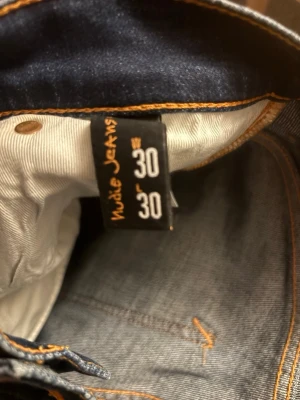 Nudie jeans Mörkblå slim fit - Snygga mörkblå jeans med slim fit passform och klassiska kontrastsömmar. Jeansen har två bakfickor med dekorativ söm och en läderpatch i midjan. Perfekta för dig som gillar en stilren och enkel look.